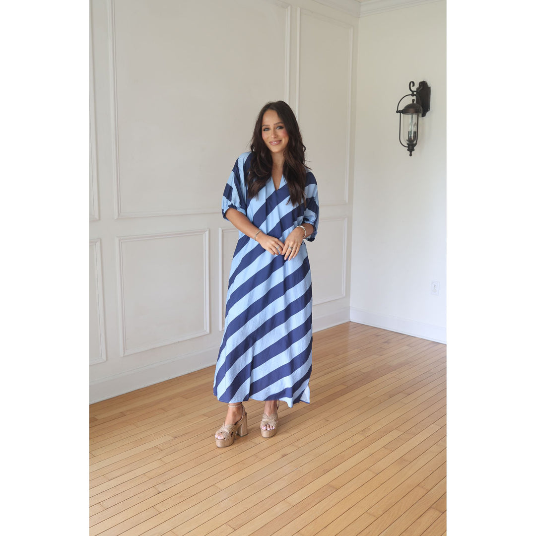 Woman wearing a blue and white striped dress standing in a room with wooden flooring and white walls.