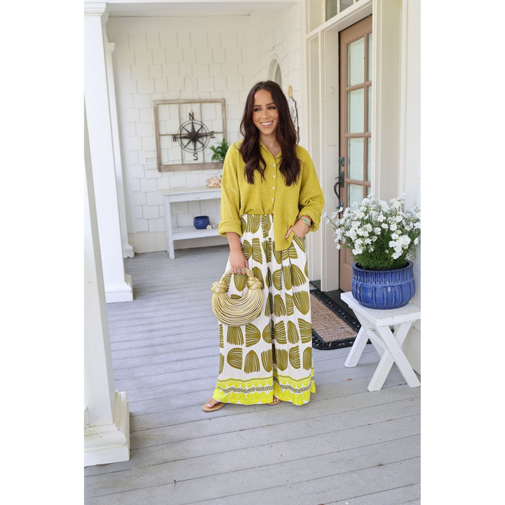 Woman in yellow top and patterned pants standing on a porch with flowers and decor.