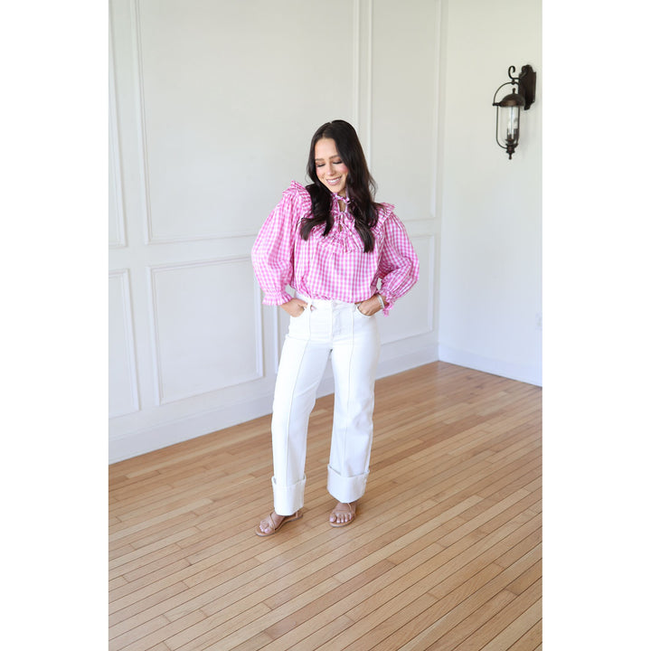 Woman wearing a pink checkered blouse and white pants standing in a room with wooden flooring and white walls.