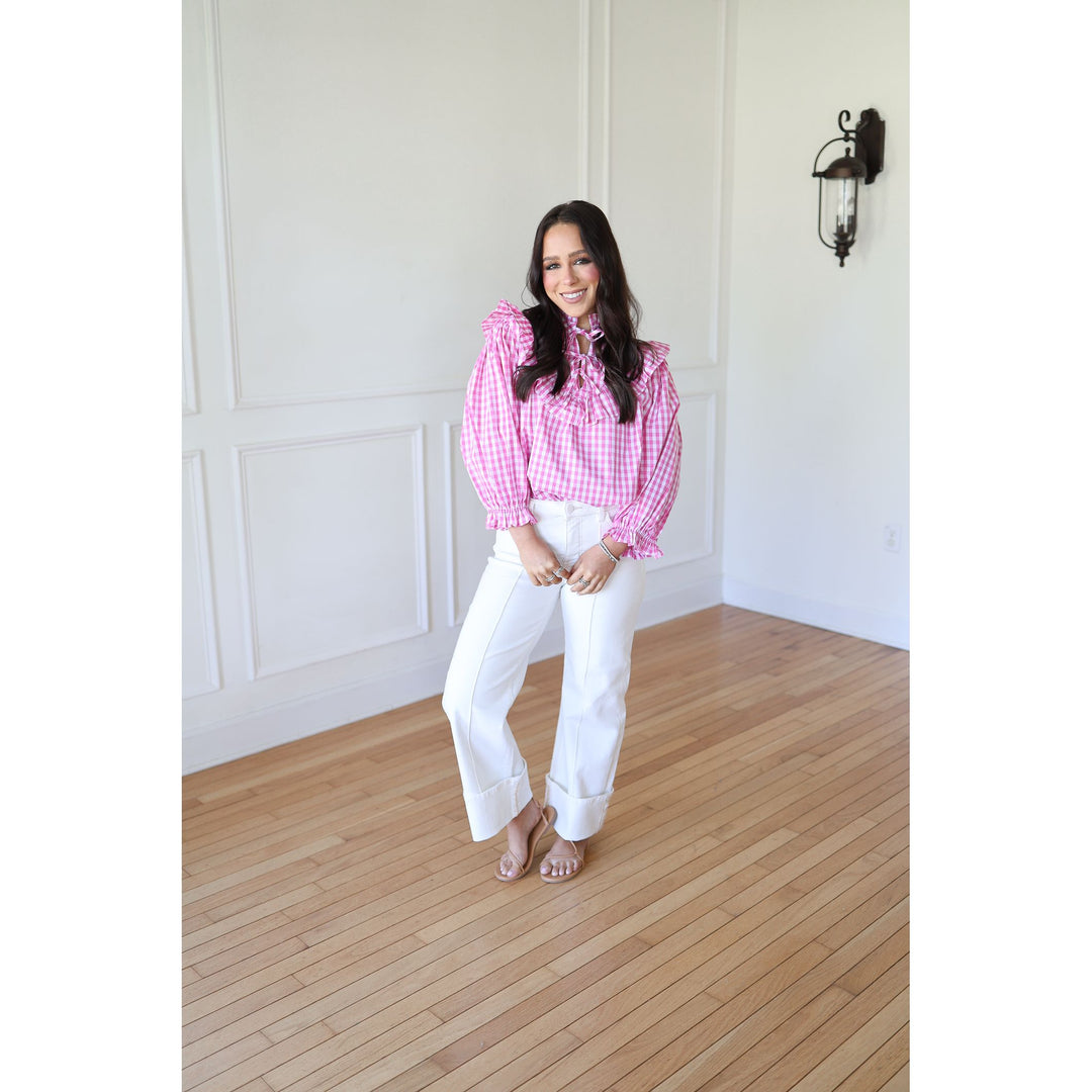 Woman wearing a pink checkered blouse and white pants standing in a room with wooden flooring and white walls.