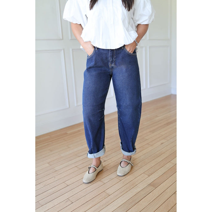 Person wearing a white blouse and blue jeans standing on a wooden floor with a white paneled wall in the background.