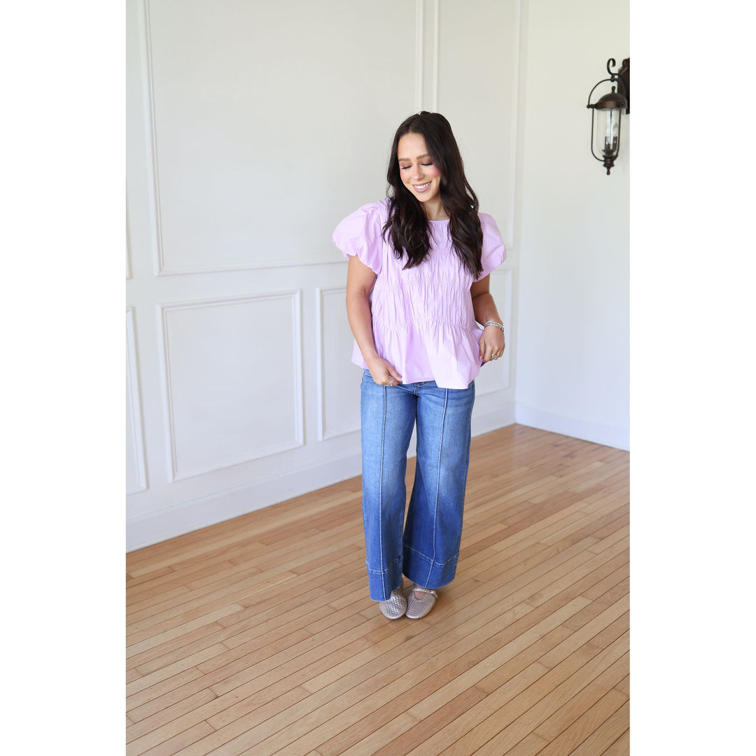 Woman wearing a light purple blouse and blue jeans standing in a room with wooden flooring and white walls.