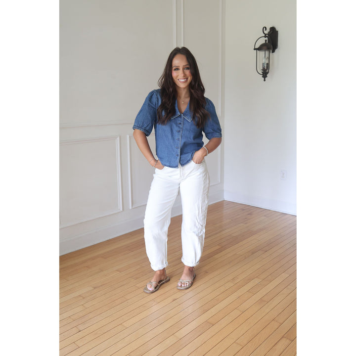 Woman wearing a blue shirt and white pants standing in a room with wooden flooring and white walls.