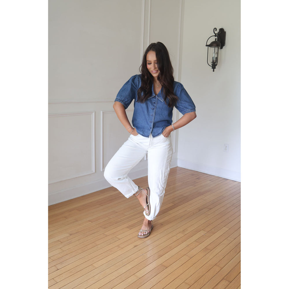 Woman wearing a blue shirt and white pants standing in a room with wooden flooring and white walls.