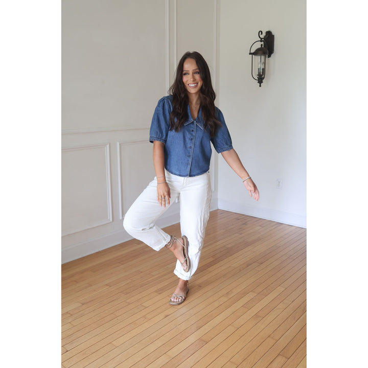 Woman wearing a blue denim shirt and white pants in a room with wooden flooring and white walls.