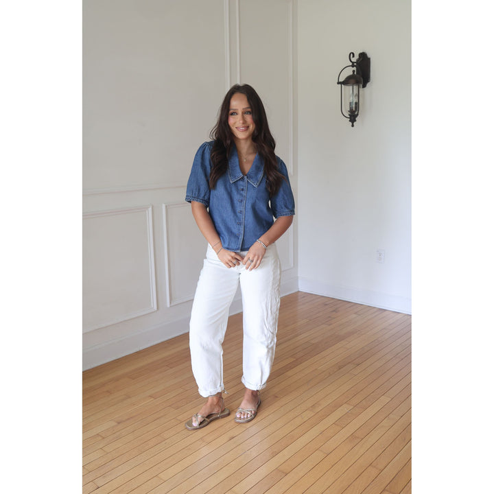Woman wearing a blue denim shirt and white pants standing in a room with wooden flooring and white walls.