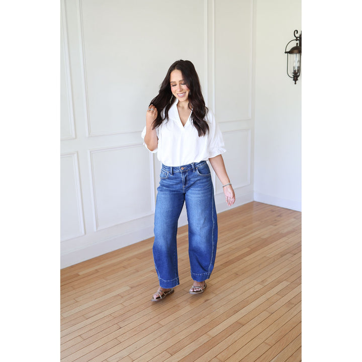 Woman wearing a white blouse and blue jeans standing in a room with wooden flooring and white walls.