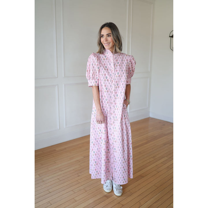 Woman wearing a pink floral dress standing in a room with wooden flooring and white walls.