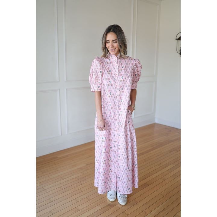 Woman wearing a pink floral dress standing in a room with wooden flooring and white walls.