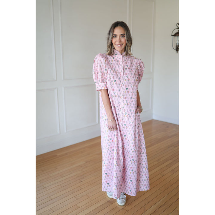Woman wearing a pink floral dress standing in a room with wooden flooring and white walls.