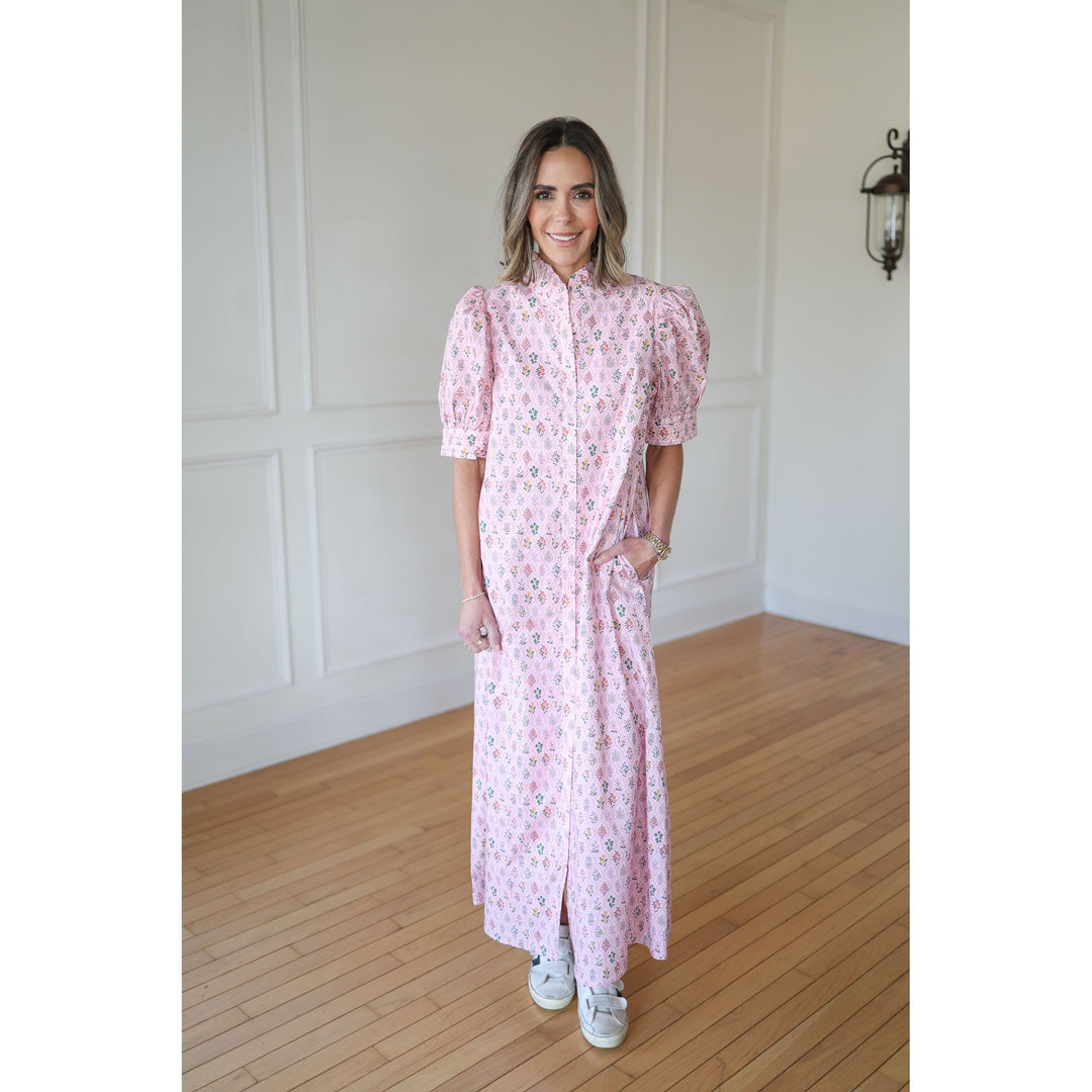 Woman wearing a pink floral dress standing in a room with wooden flooring and white walls.