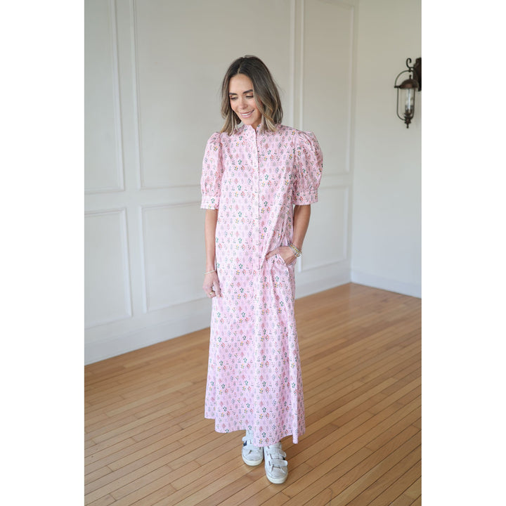 Woman wearing a pink floral dress standing in a room with wooden flooring and white walls.