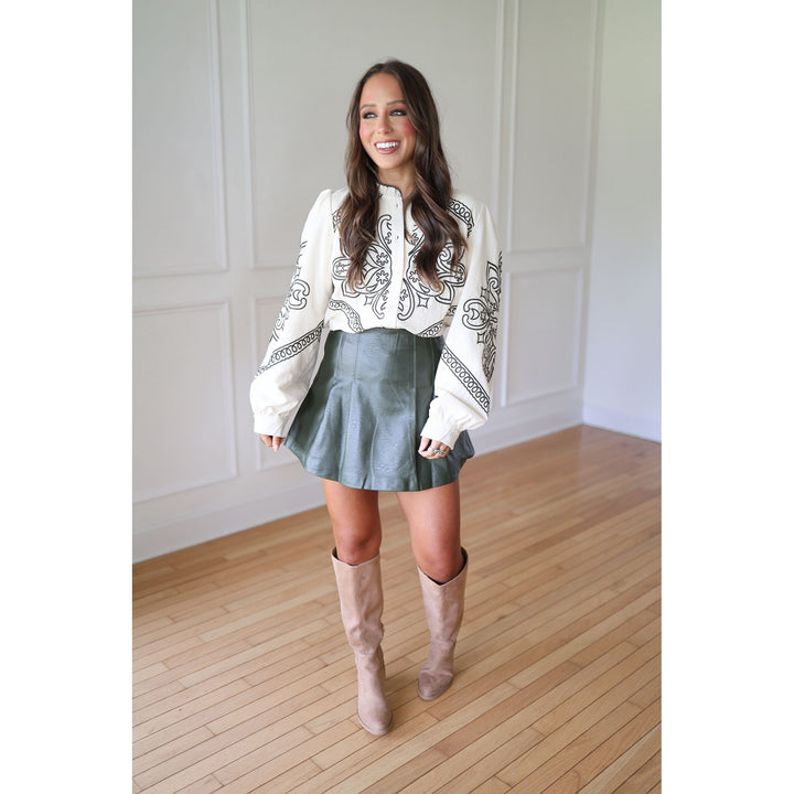 Woman wearing a white blouse with green patterns, green skirt, and beige knee-high boots 