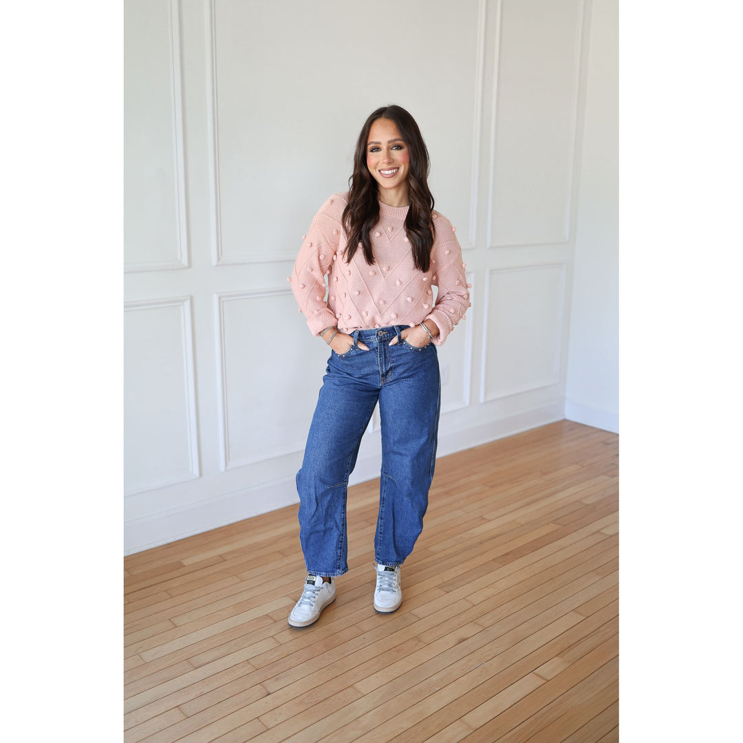 Woman wearing a pink sweater and blue jeans standing in a room with wooden flooring and white paneled walls.
