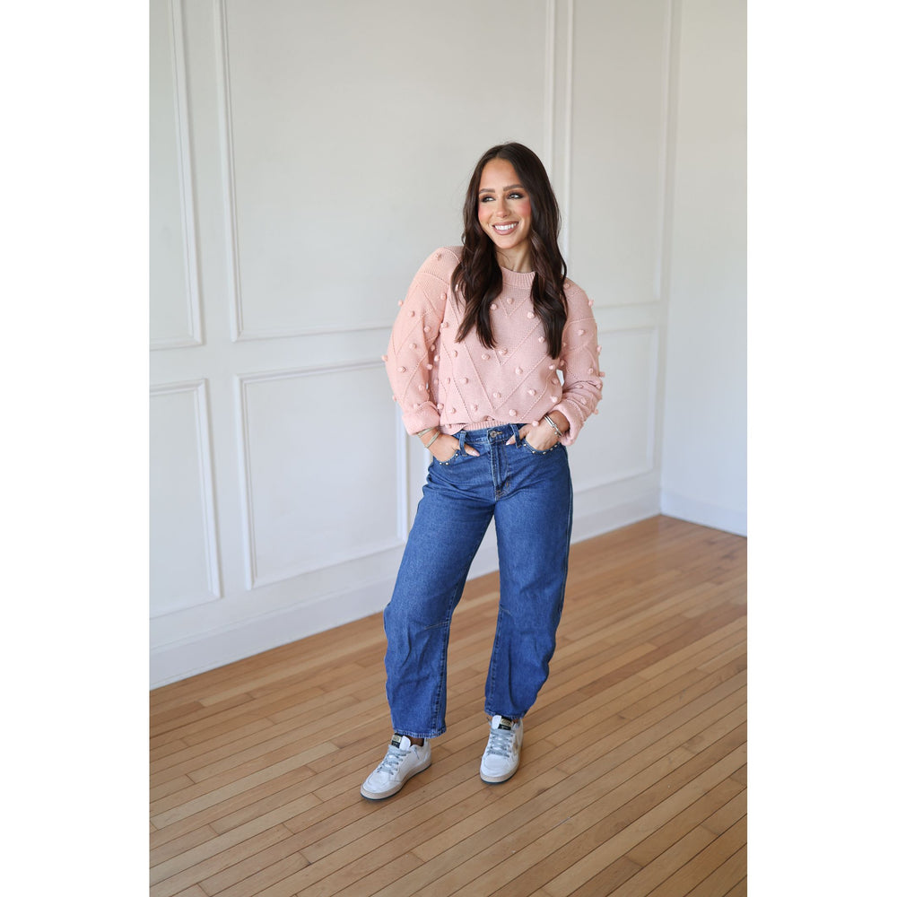 Woman wearing a pink sweater with white polka dots and blue jeans standing in a room with wooden flooring and white walls.