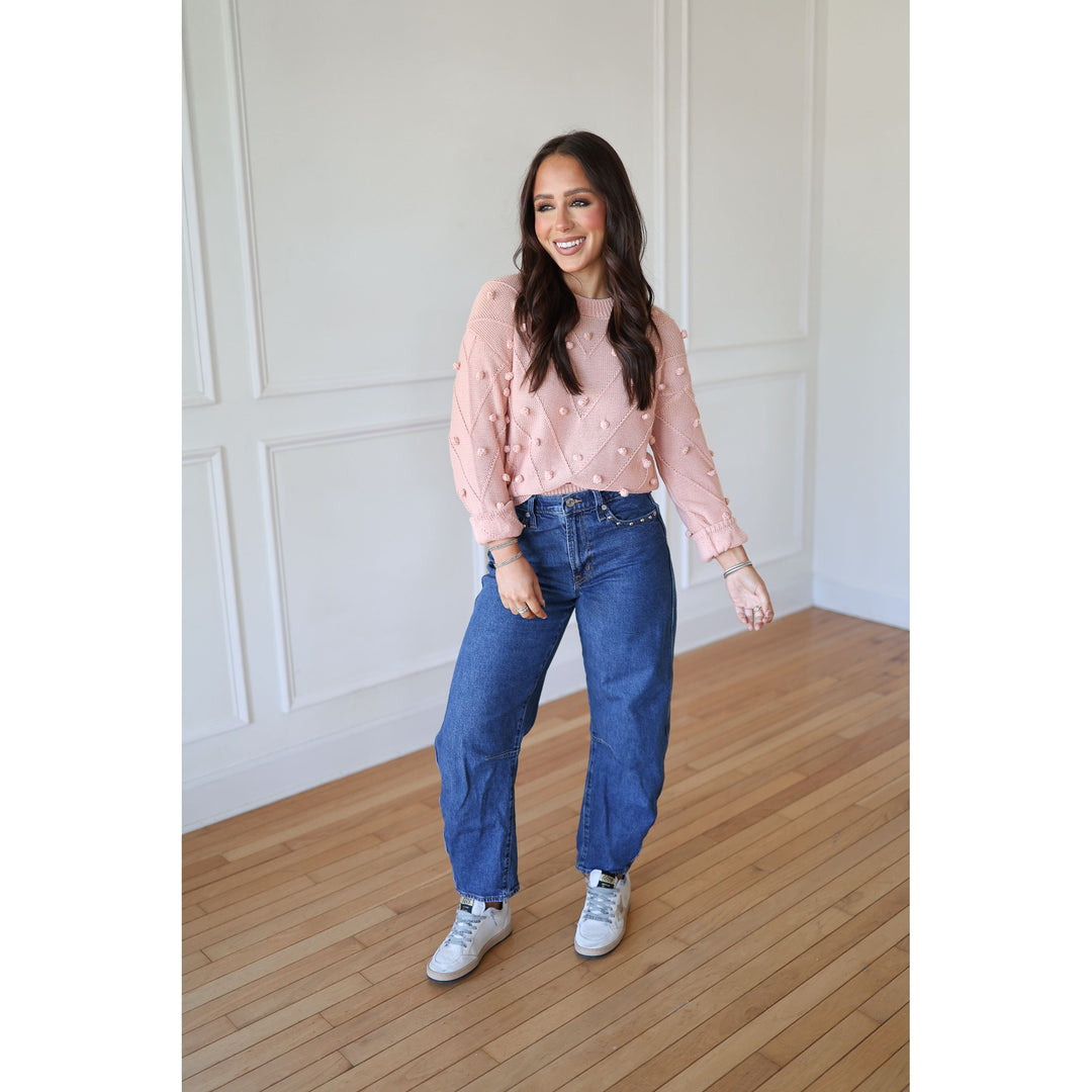 Woman wearing a pink sweater with white polka dots and blue jeans standing in a room with wooden flooring and white paneled walls.