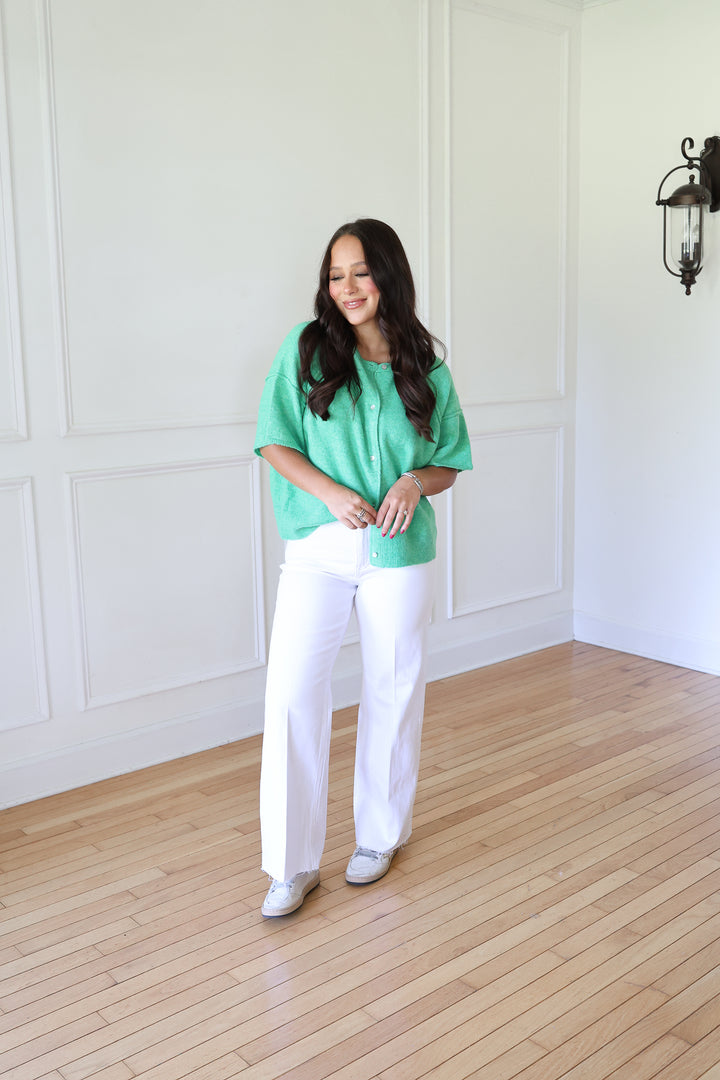 A woman stands in a bright room with white paneled walls and light wood floors, wearing a mint green short-sleeve button-up top, white wide-leg pants, and white sneakers.