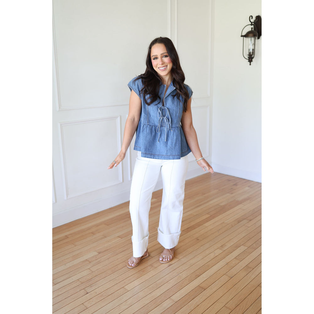 Woman wearing a denim top and white pants standing in a room with wooden flooring and white walls.
