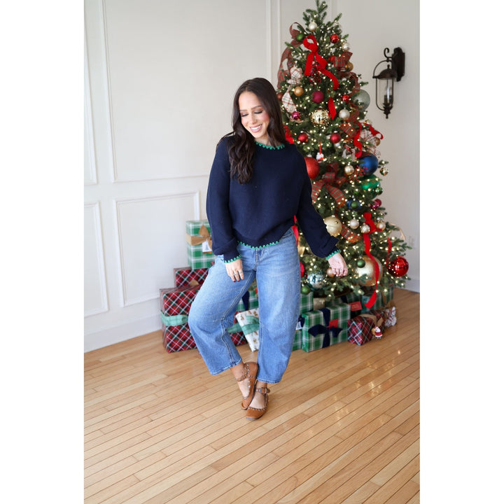 Woman in a navy sweater and jeans standing in front of a decorated Christmas tree with presents underneath.
