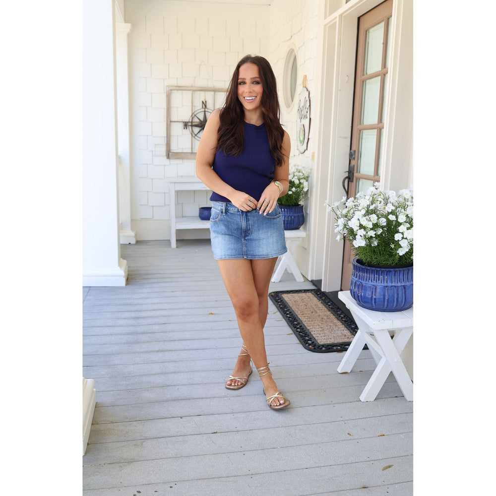 Woman standing on a porch wearing a blue top and denim shorts, with a white door and plants in the background.