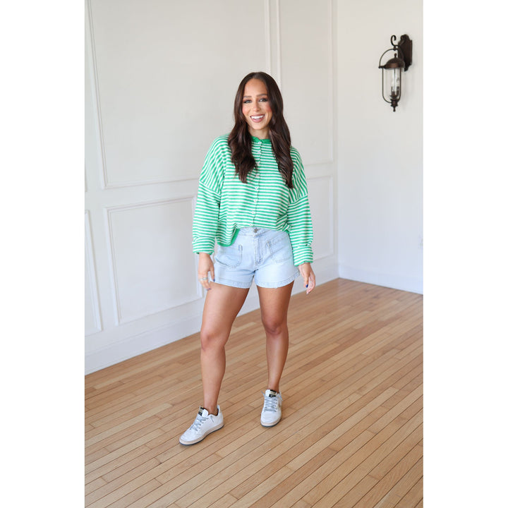 Woman wearing a green striped sweater, denim shorts, and white sneakers in a room with wooden flooring and white walls.