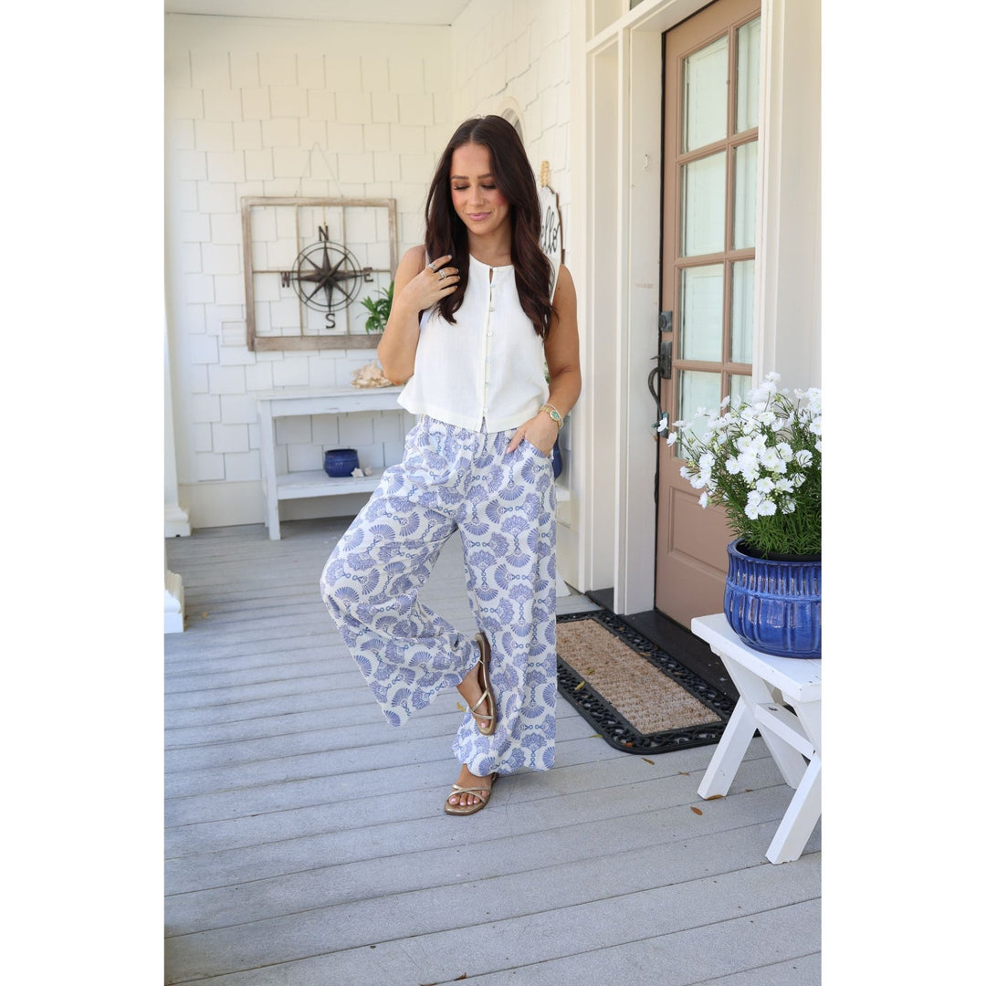 Woman in white sleeveless top and patterned pants standing on a wooden deck with a white house in the background.