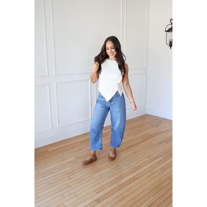 Woman in a white sleeveless top and blue jeans standing on a wooden floor.