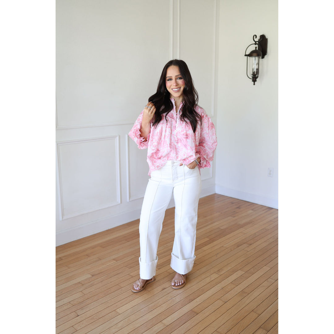 Woman wearing a pink blouse and white pants standing in a room with wooden flooring and white walls.