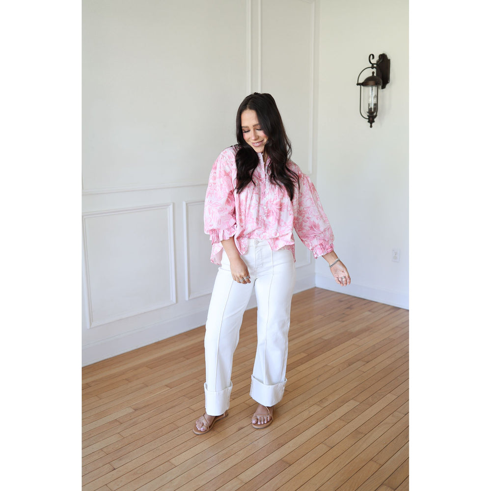 Woman wearing a pink blouse and white pants in a room with wooden flooring and white walls.