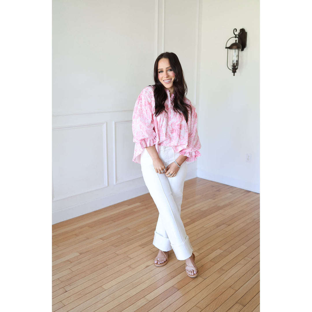 Woman wearing a pink blouse and white pants standing in a room with wooden flooring and white walls.