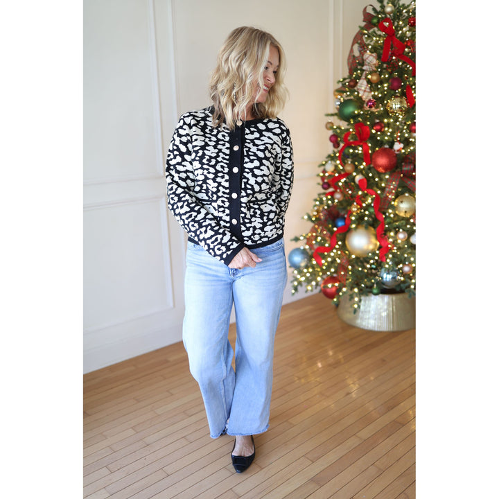 Woman wearing a black and white patterned cardigan and light blue jeans standing in front of a decorated Christmas tree.