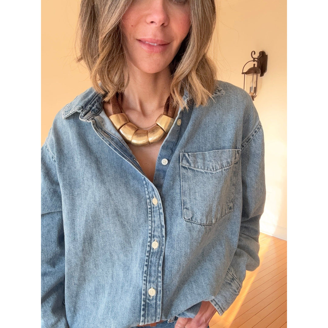 Person wearing a light blue denim shirt with a gold necklace against a neutral background