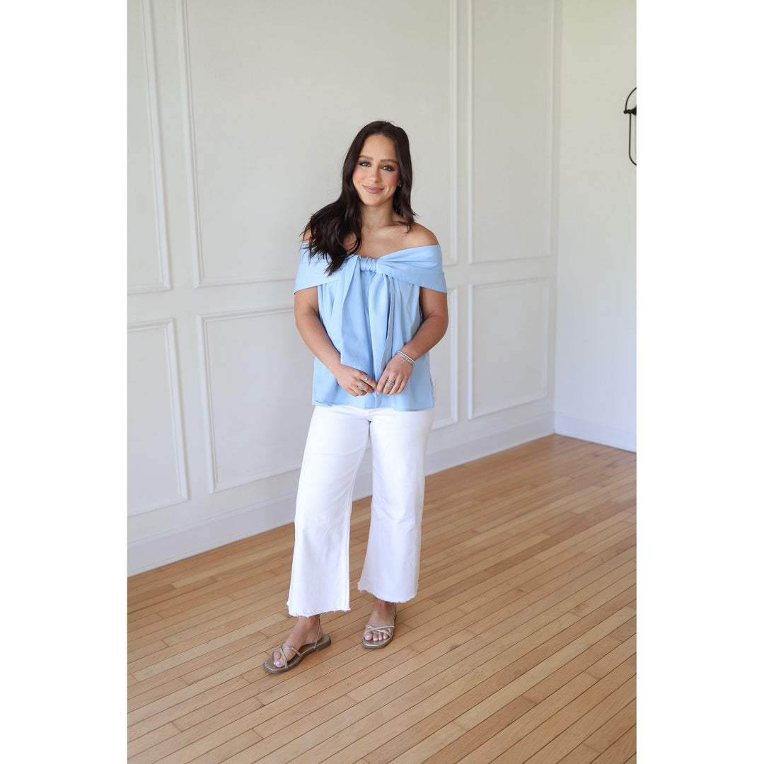 Woman wearing a light blue off-shoulder top and white pants standing in a room with wooden flooring and white paneled walls.