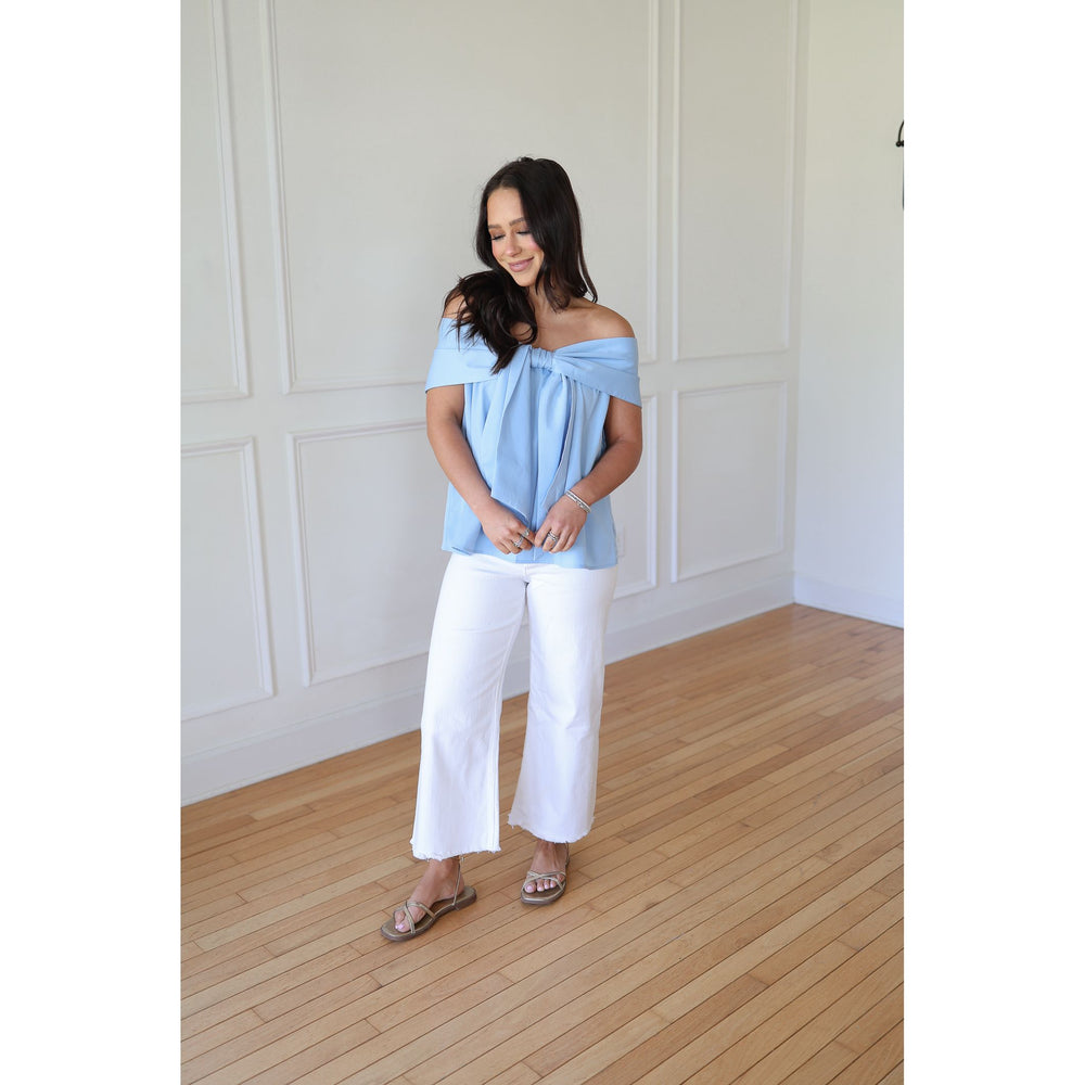 Woman wearing a light blue off-shoulder top and white pants in a room with wooden flooring and white walls.