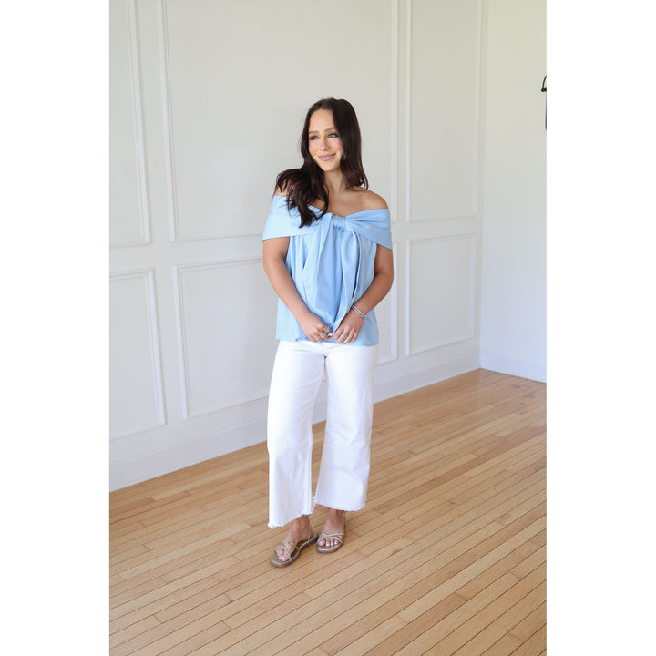 Woman wearing a light blue off-shoulder top and white pants in a room with wooden flooring and white walls.