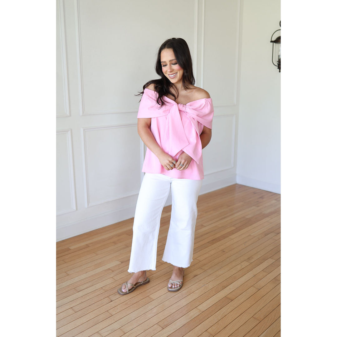 Woman wearing a pink off-shoulder top and white pants in a room with wooden flooring.