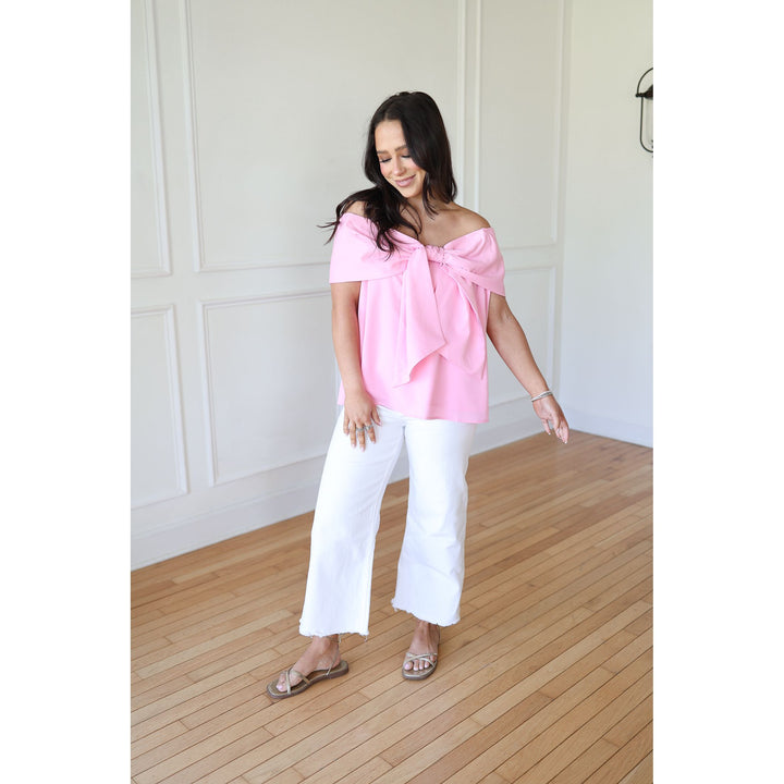 Woman wearing a pink off-shoulder top and white pants in a room with wooden flooring.