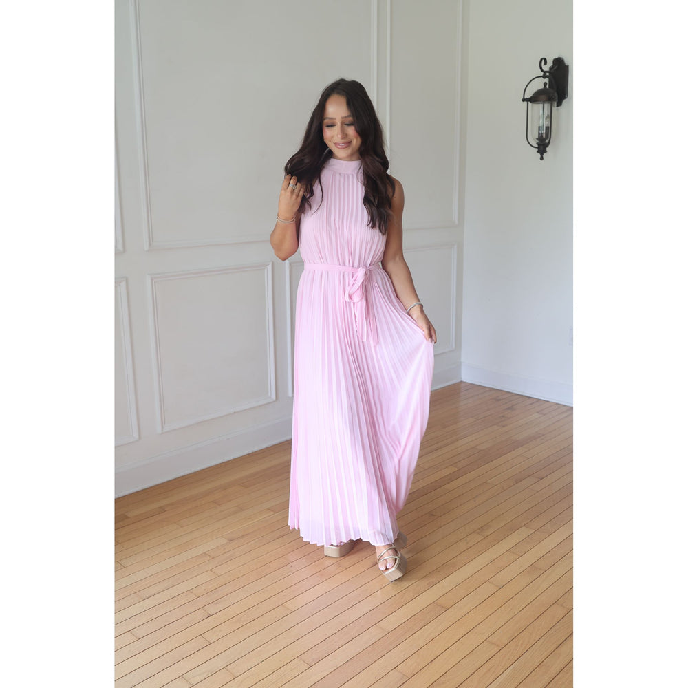 Woman wearing a pink pleated dress in a room with wooden flooring and white walls.