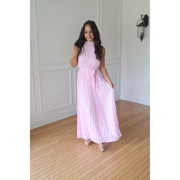 Woman in a pink pleated dress standing in a room with wooden flooring and white walls.
