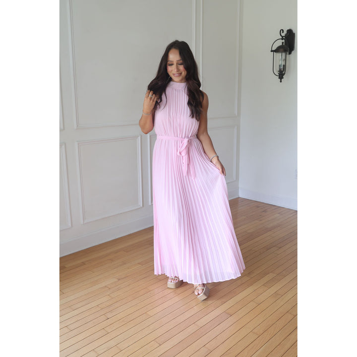Woman wearing a pink pleated dress standing in a room with wooden flooring and white walls.
