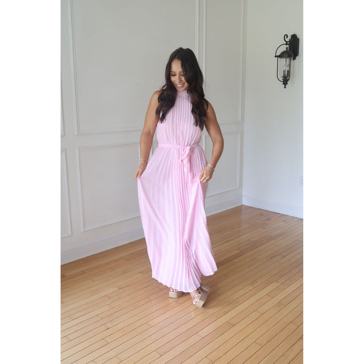 Woman wearing a pink pleated dress in a room with wooden flooring and white walls.