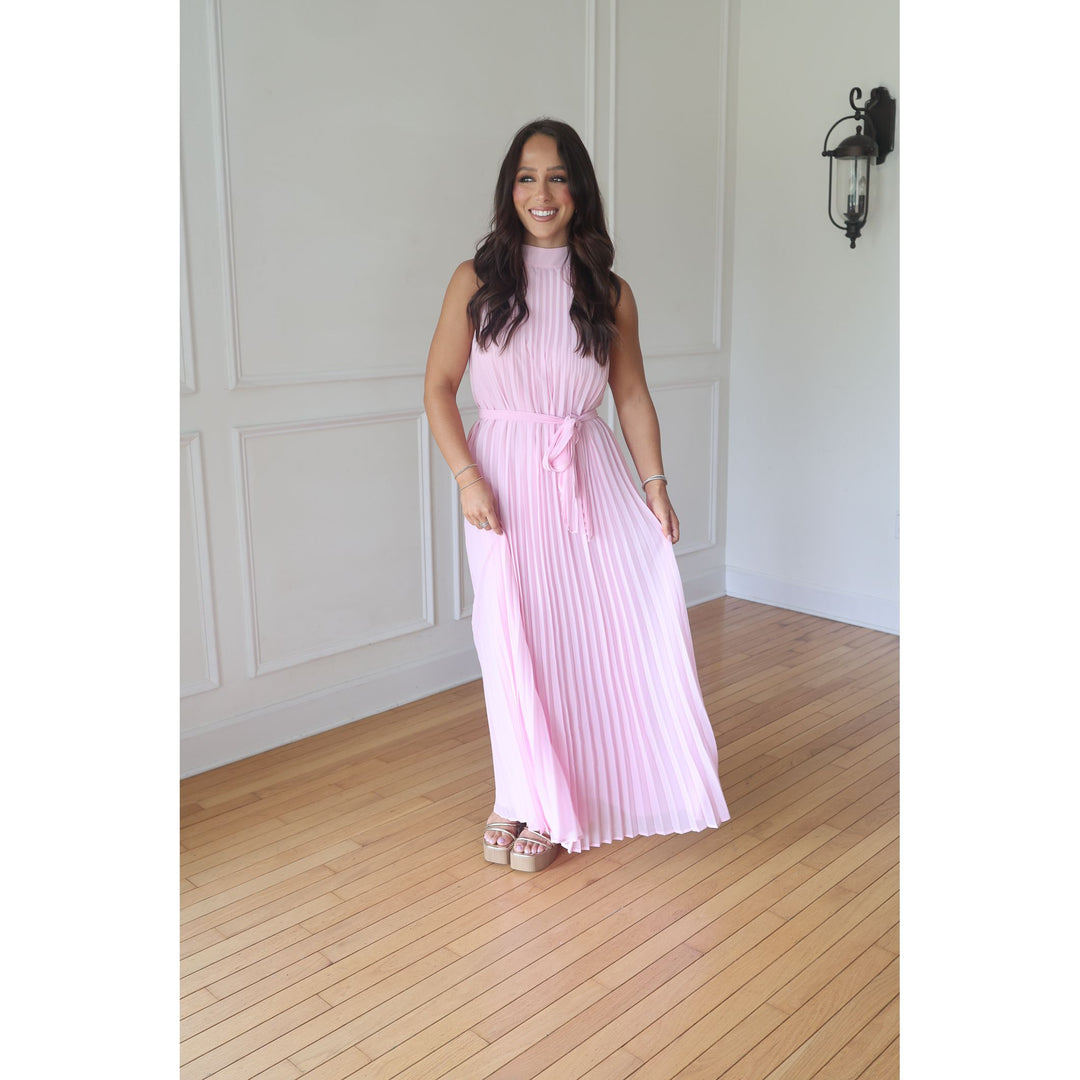 Woman wearing a pink pleated dress standing in a room with wooden flooring and white walls.