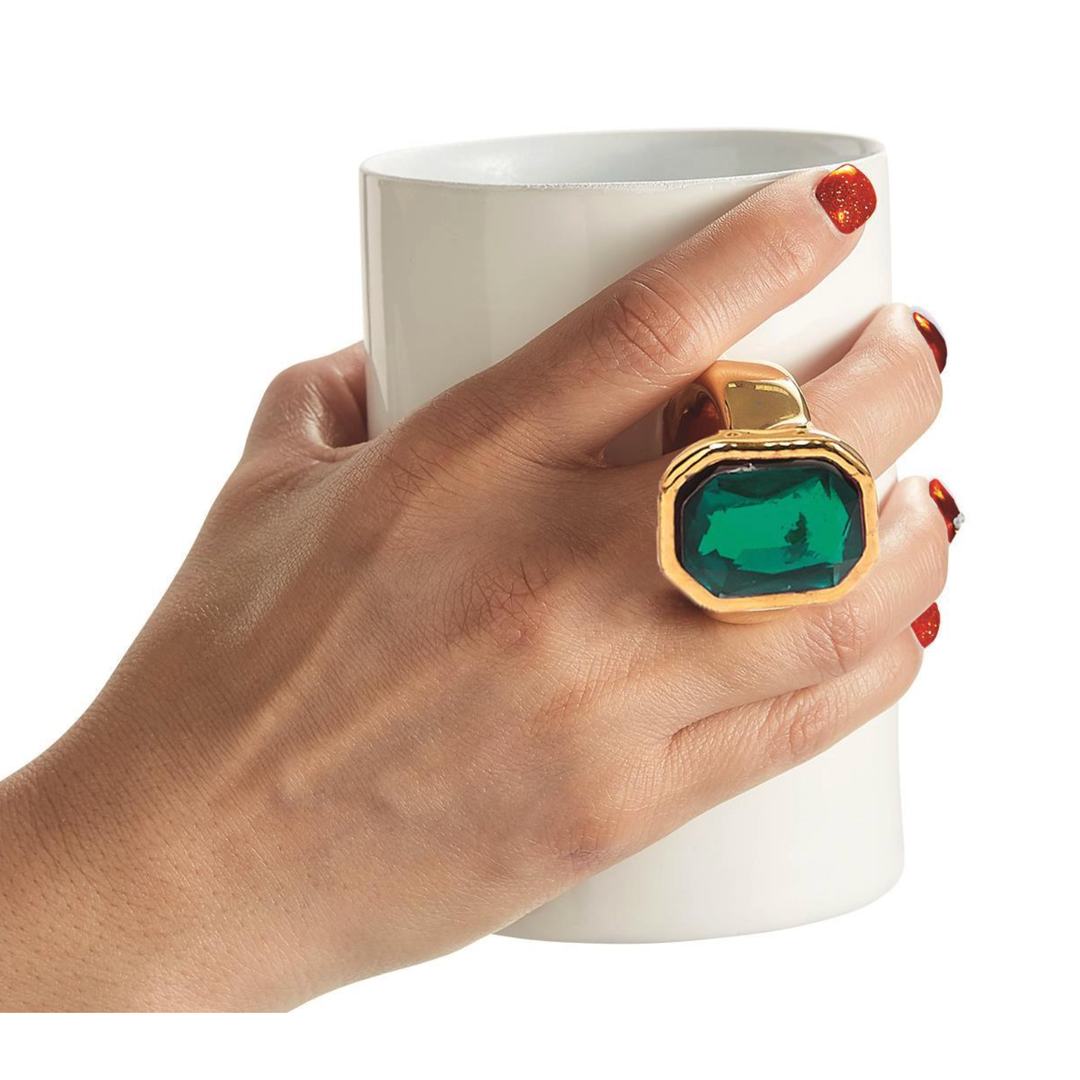 Hand wearing a gold ring with a green gemstone holding a white mug.