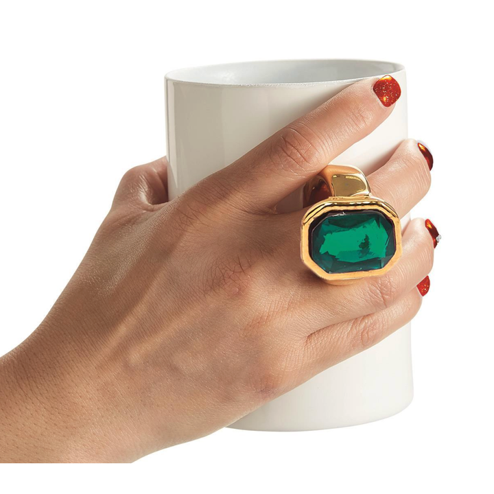 Hand wearing a gold ring with a green gemstone holding a white mug.