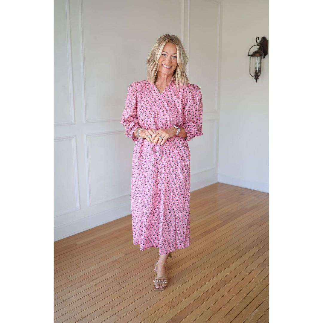 Woman wearing a pink patterned dress standing in a room with wooden flooring and white walls.