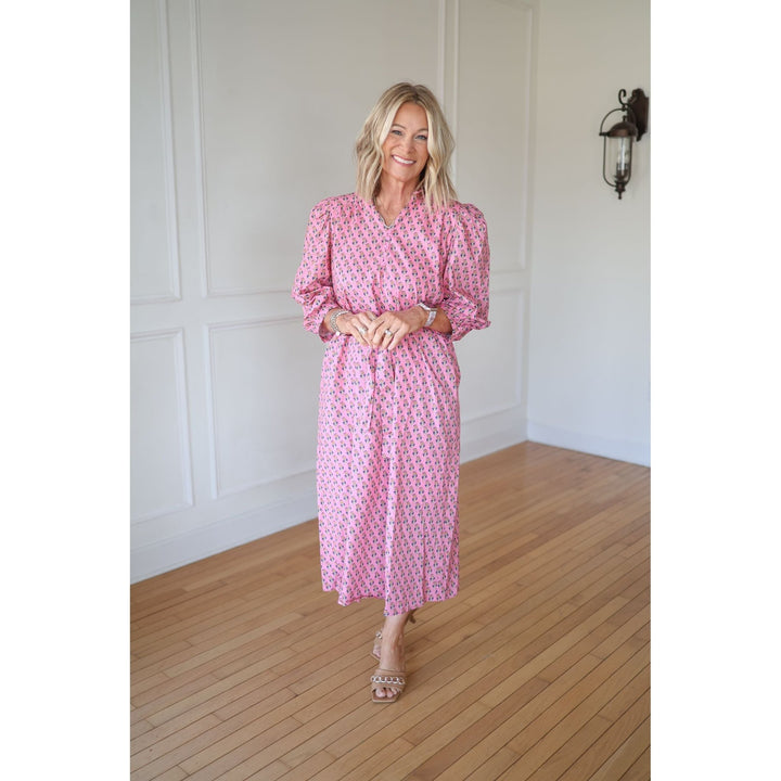 Woman wearing a pink patterned dress standing in a room with wooden flooring and white walls.