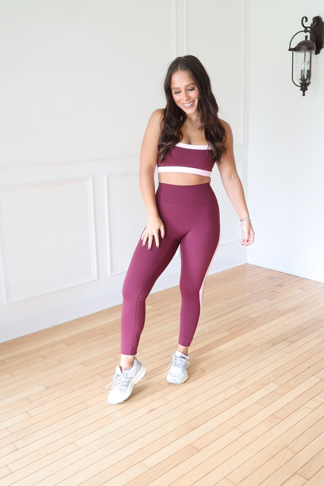 A woman modeling a matching burgundy activewear set consisting of a sports bra and high-waisted leggings with white side stripes