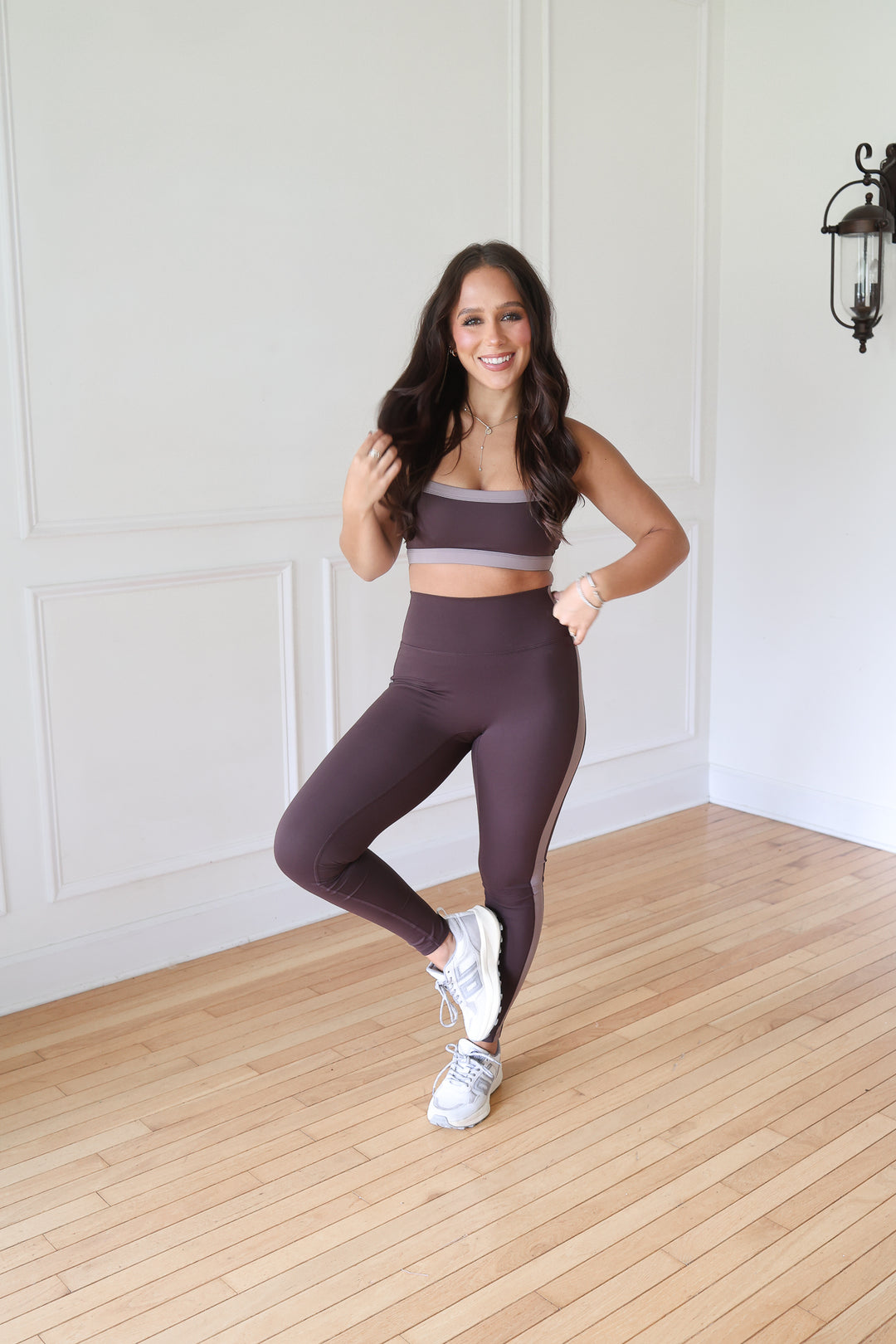 A woman posing in a matching two-piece activewear set consisting of a dark plum purple sports bra and high-waisted leggings with white sneakers