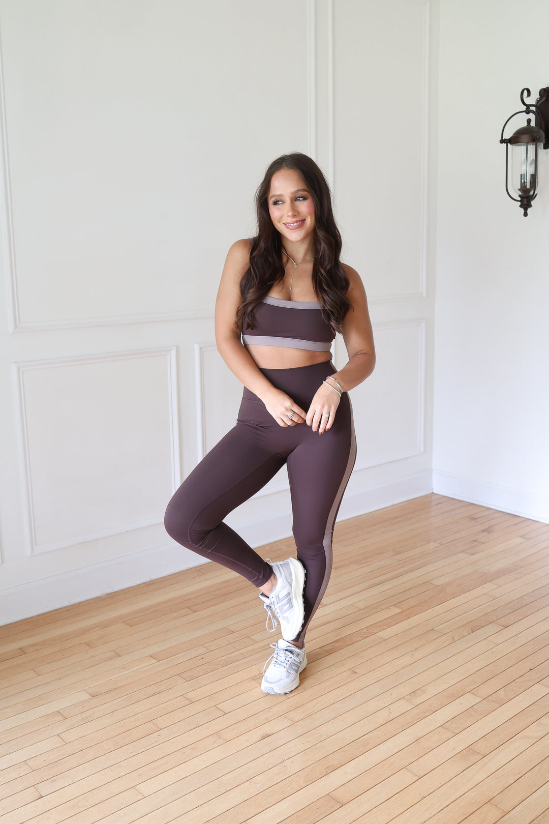 A woman posing in a matching athletic set consisting of a dark plum purple sports bra and high-waisted leggings with a thin light gray side stripe, paired with white sneakers