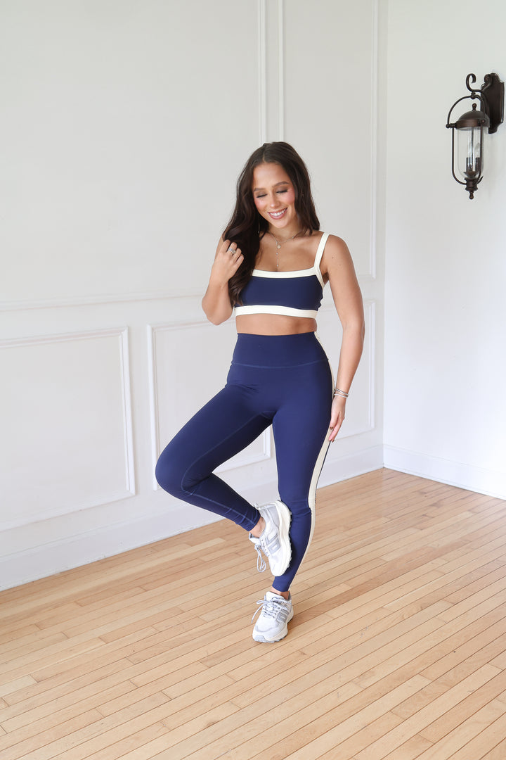 A woman modeling a matching navy blue activewear set consisting of a sports bra and high-waisted leggings with white contrast trim